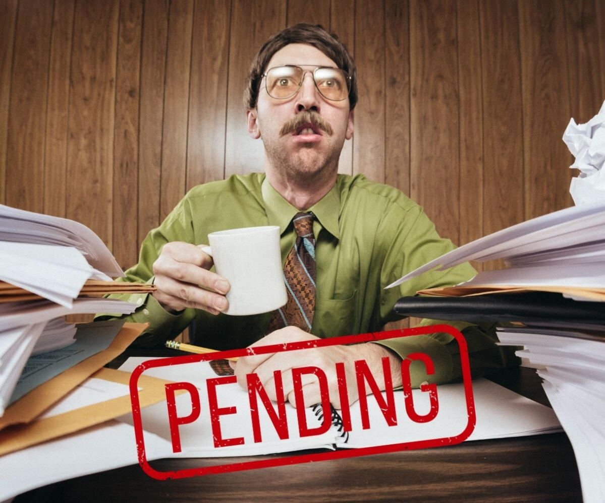 An exhausted office worker smiles nervously at his desk, surrounded by stacks of paperwork, holding a cup of coffee. The word "PENDING" is stamped across the image in red.